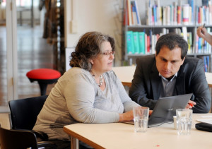 Foto mit einer Frau und einem Mann, die am Tisch sitzen und auf einen Laptop gucken