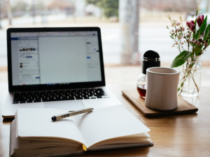 Foto mit einem Laptop, ein Buch darauf ein Stift ist im Vordergrund und eine Tasse steht auf dem Schreibtisch