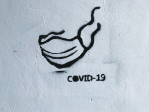 Foto mit einer Hauswand welche besprüht wurde mit der Aufschrift: COVID-19 und eine gemalte Maske danehben.