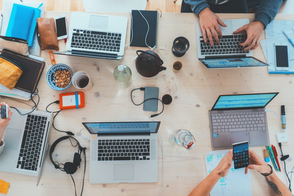 Das Foto eines Tisches von oben. Darauf: viele aufgeklappte Laptops und Geräte. Zudem sieht man Hände.