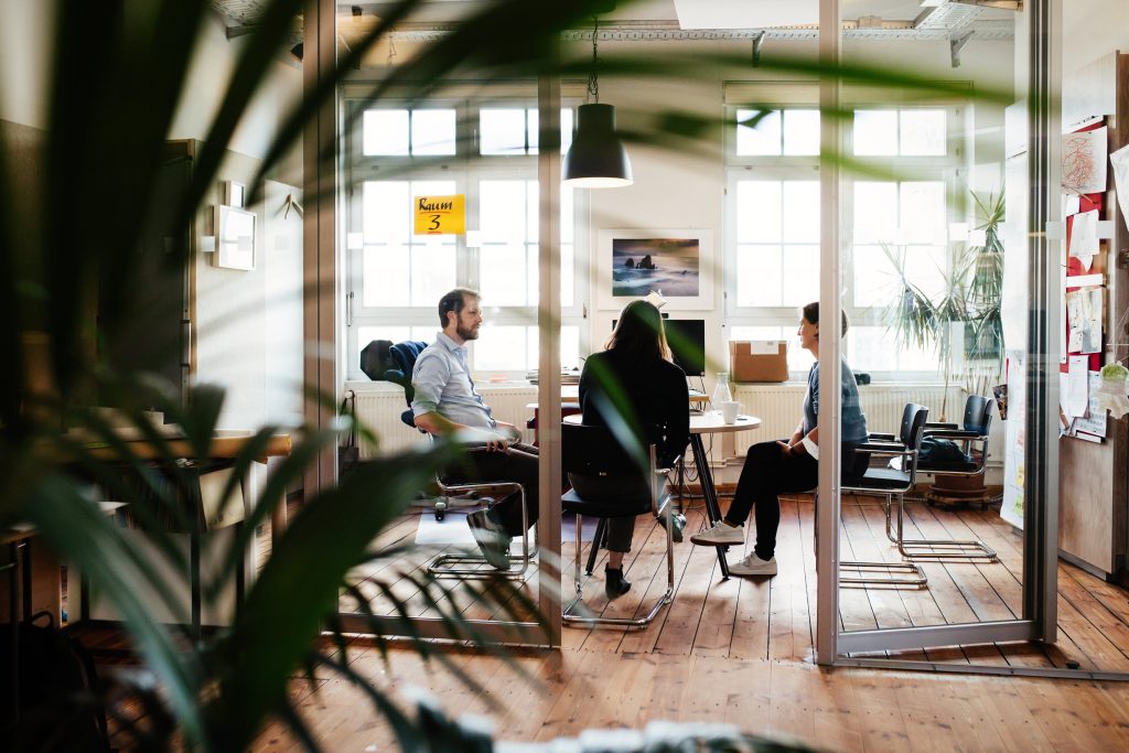 Zwei Frauen und ein Mann sitzen in einem Büro um einen runden Tisch und unterhalten sich. Zwischen Fotograf und Persoenen ist eine Glastür, und die spitzen einer Pflanze sind undeutlich im Vordergrund.